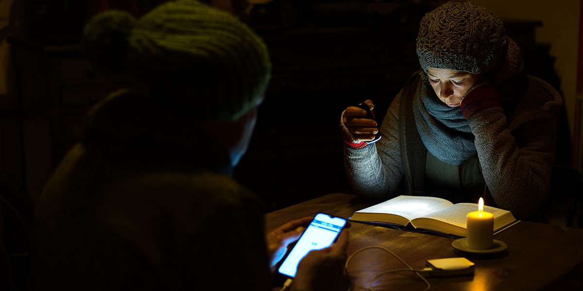 Kobieta czytająca książkę w blasku latarki i kobieta używająca telefonu podłączonego do powerbanka w ciemnym pokoju